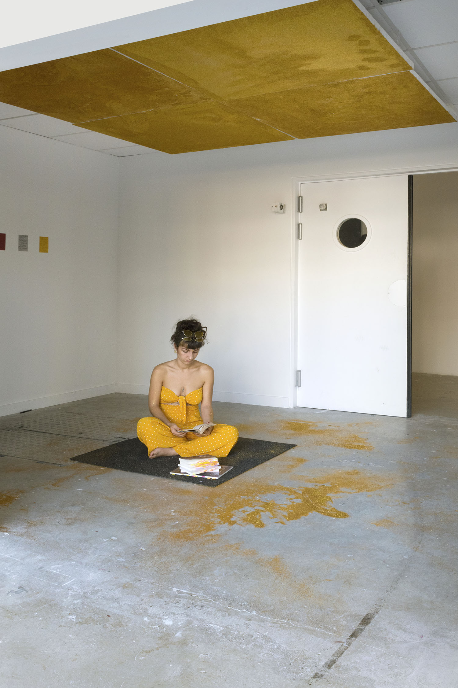 une femme lit sur un carrée de moquette. Au-dessus d'elle, 4 écrans recouverts de paillettes dorées sont installés. Des paillettes tombent sur elle et se répendent au sol.